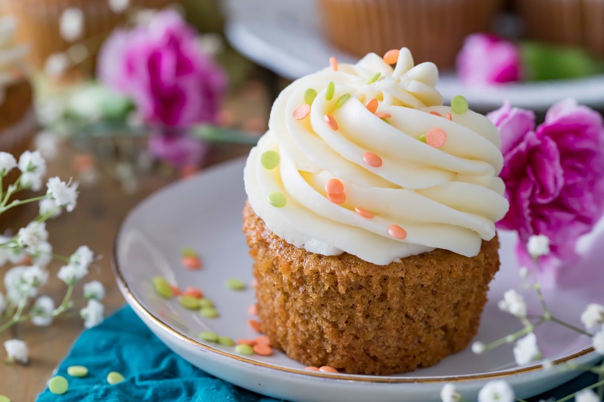 Soft, fluffy, and moist carrot cake cupcakes! Made completely from scratch with real grated carrots and the option to add nuts or raisins (I would never, but you do you...), these cupcakes are always a hit! Make sure to top them off with the absolute best cream cheese frosting. Carrot Cake Cupcake on a plate, surrounded by flowers