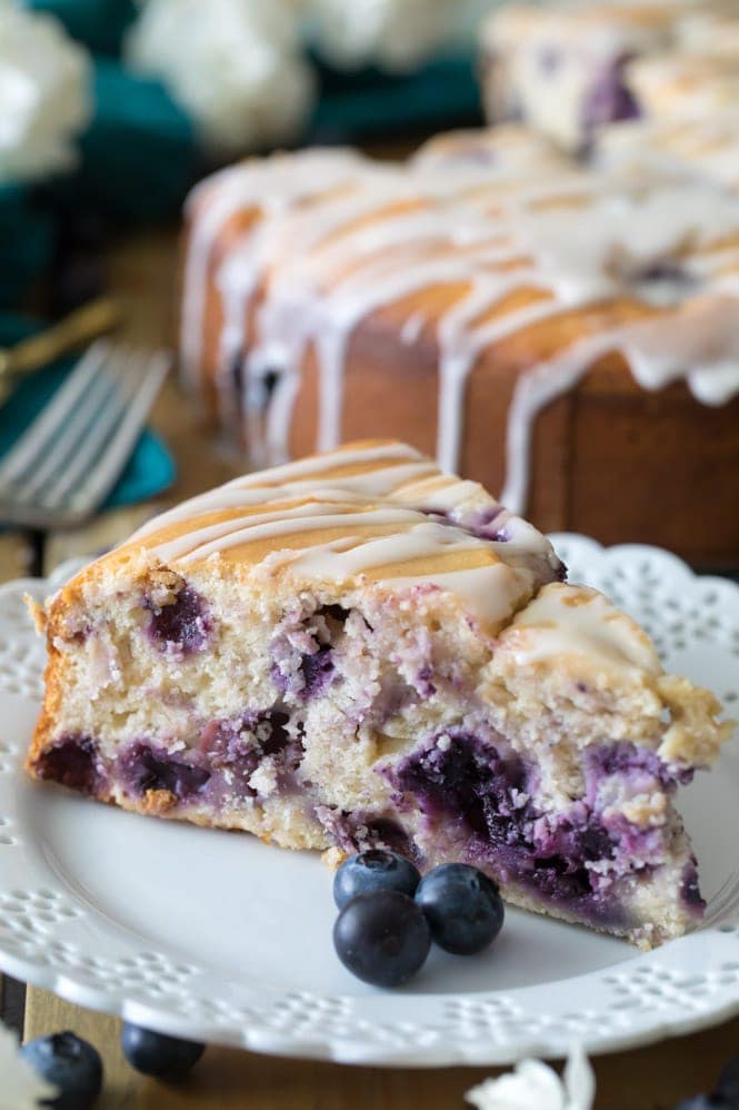 How to make a BLUEBERRY CAKE! Packed full of fresh blueberries and topped off with a lemon glaze, you're going to want to save the recipe for this one! #blueberrycake #cake #recipe #baking #blueberries slice of blueberry cake on a white plate