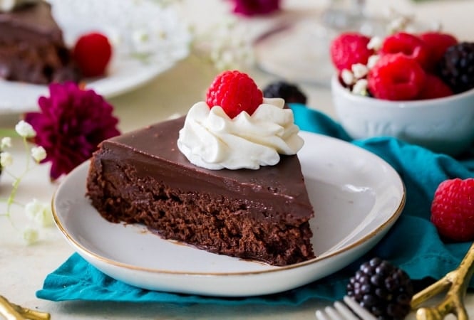A rich, dense, & supremely fudgy FLOURLESS CHOCOLATE CAKE! #flourlesschocolatecake #glutenfree Slice of flourless chocolate cake on plate topped with berries and cream