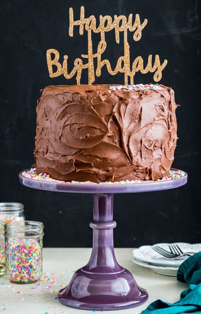 The BEST BIRTHDAY CAKE RECIPE! This vanilla cake is light and fluffy and packed full of colorful sprinkles! #birthdaycake #birthdayparty #easycakerecipe Chocolate frosted birthday cake on a purple cake stand