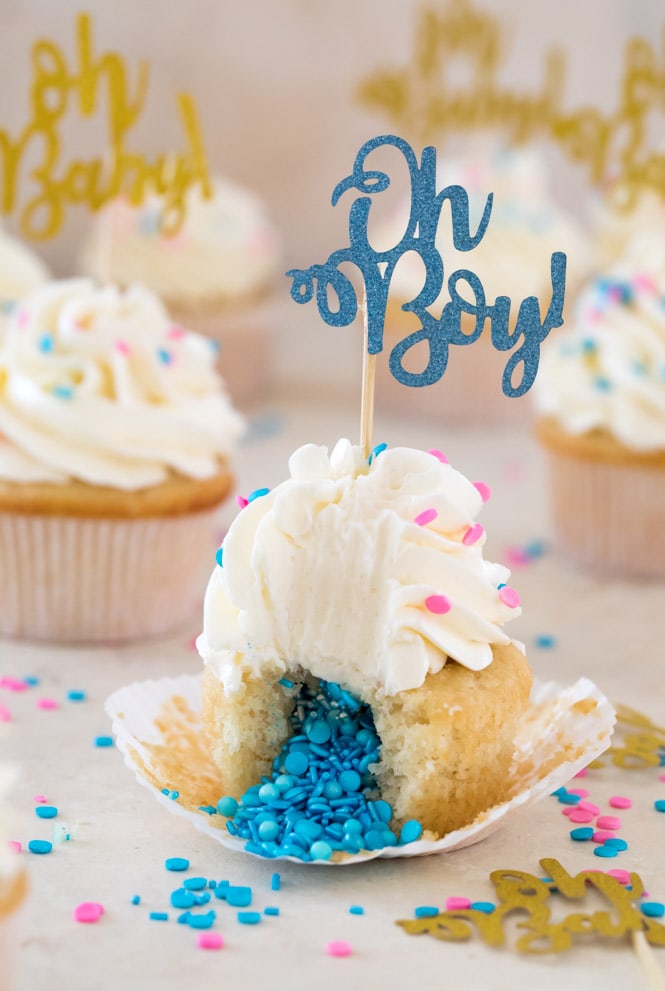 A sweet and simple recipe for Gender Reveal Cupcakes! Fill them with either pink or blue sprinkles for a fun surprise reveal #genderreveal #genderrevealcupcakes #genderrevealcake #babyshower #sugarspunrun Gender reveal cupcake cut in half with blue sprinkles pouring out of center