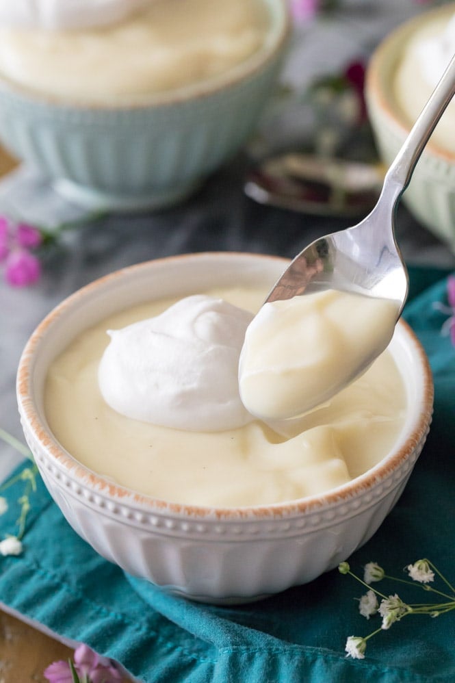 HOMEMADE VANILLA PUDDING! So easy to make with ingredients you already have on hand, no gelatin, no fancy odd ingredients! This is a great easy pudding recipe! #pudding #puddingrecipe #sugarspunrun Spoon with vanilla pudding on it, bowl of vanilla pudding topped with whipped cream in the background