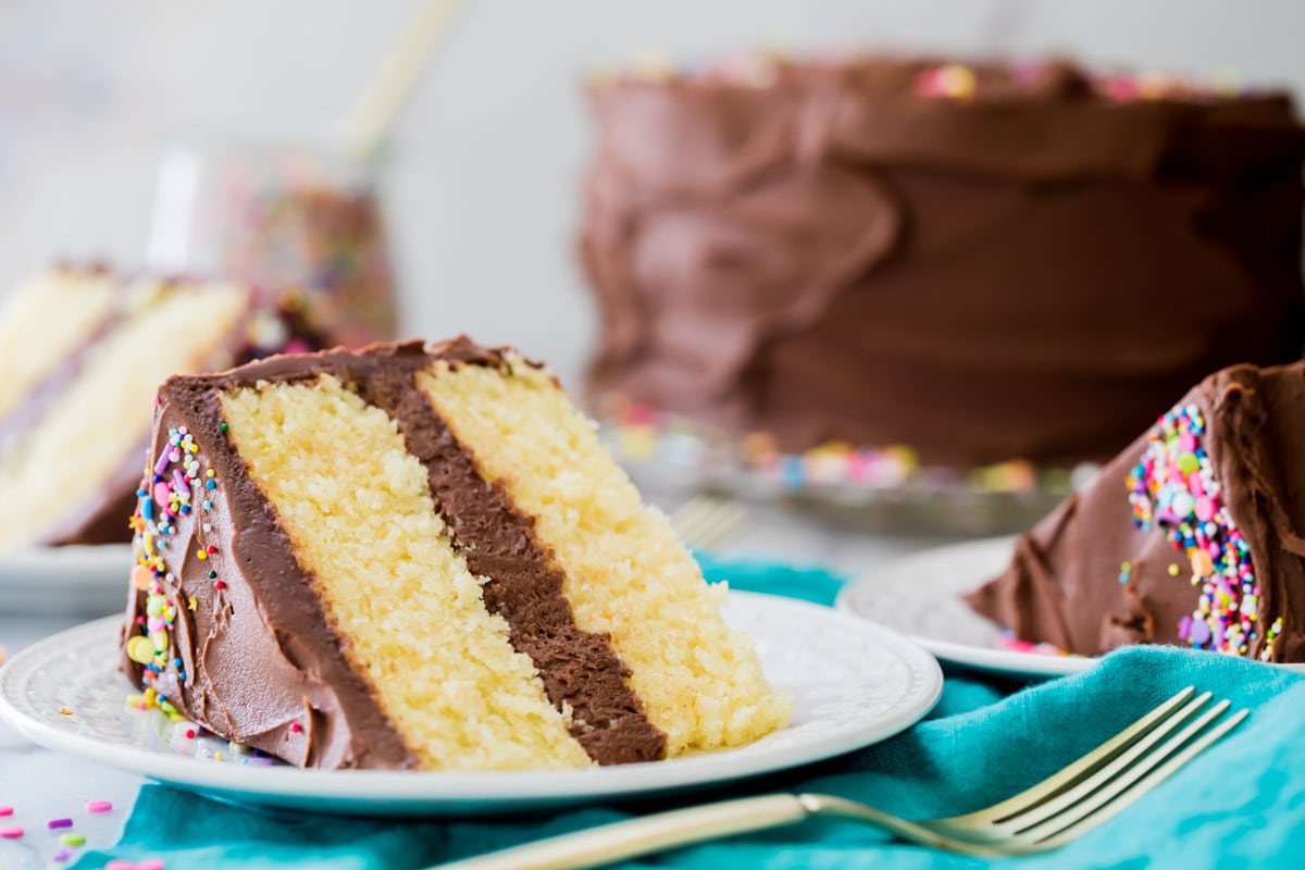 A slice of two-layer vanilla cake on a white plate with cake and other slices in background.