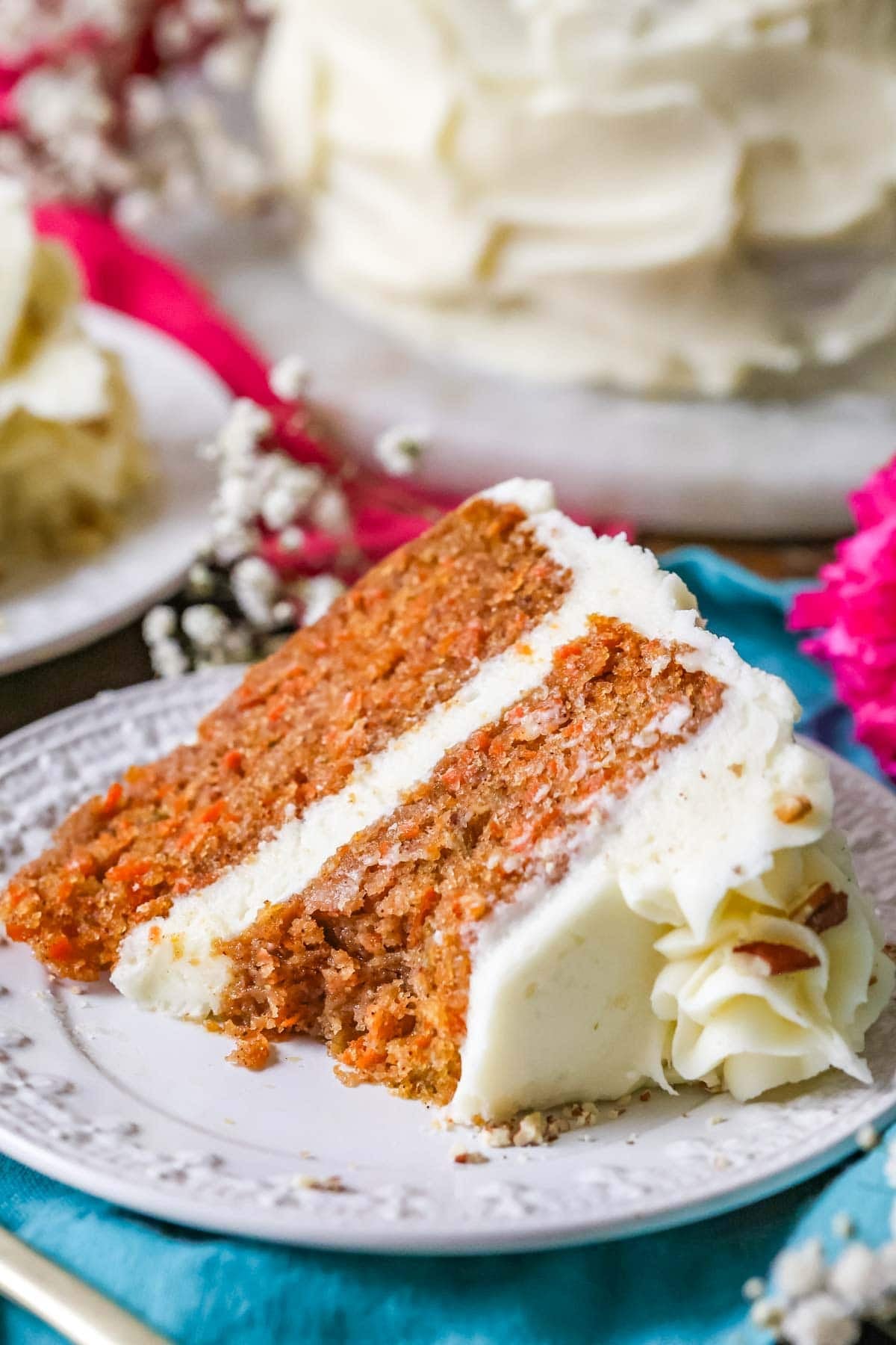 A slice of the best carrot cake recipe on a white plate with white flowers in the background. A bite missing from the slice.