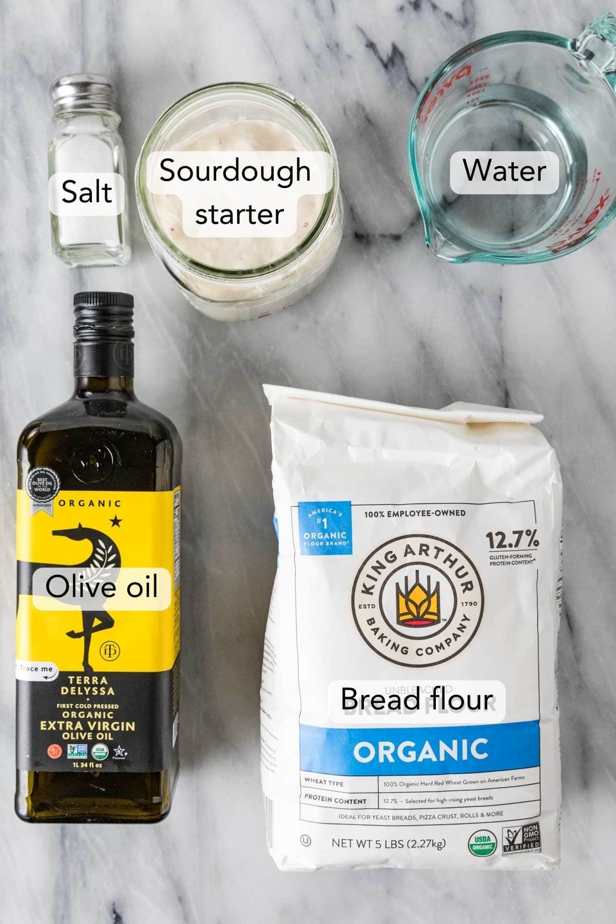 Overhead view of ingredients needed for the sourdough bread recipe including olive oil, bread flour, sourdough starter, and more.