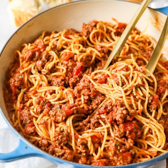 Tongs tossing spaghetti with meat sauce in a large pot.