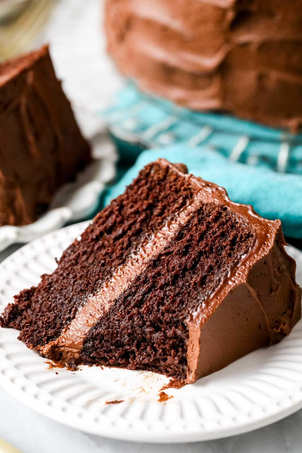 Slice of fudgy chocolate cake on a white plate with cake on crystal platter over blue cloth in background.