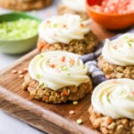 Carrot cake cookies toped with cream cheese frosting and sprinkles on a wood cutting board.