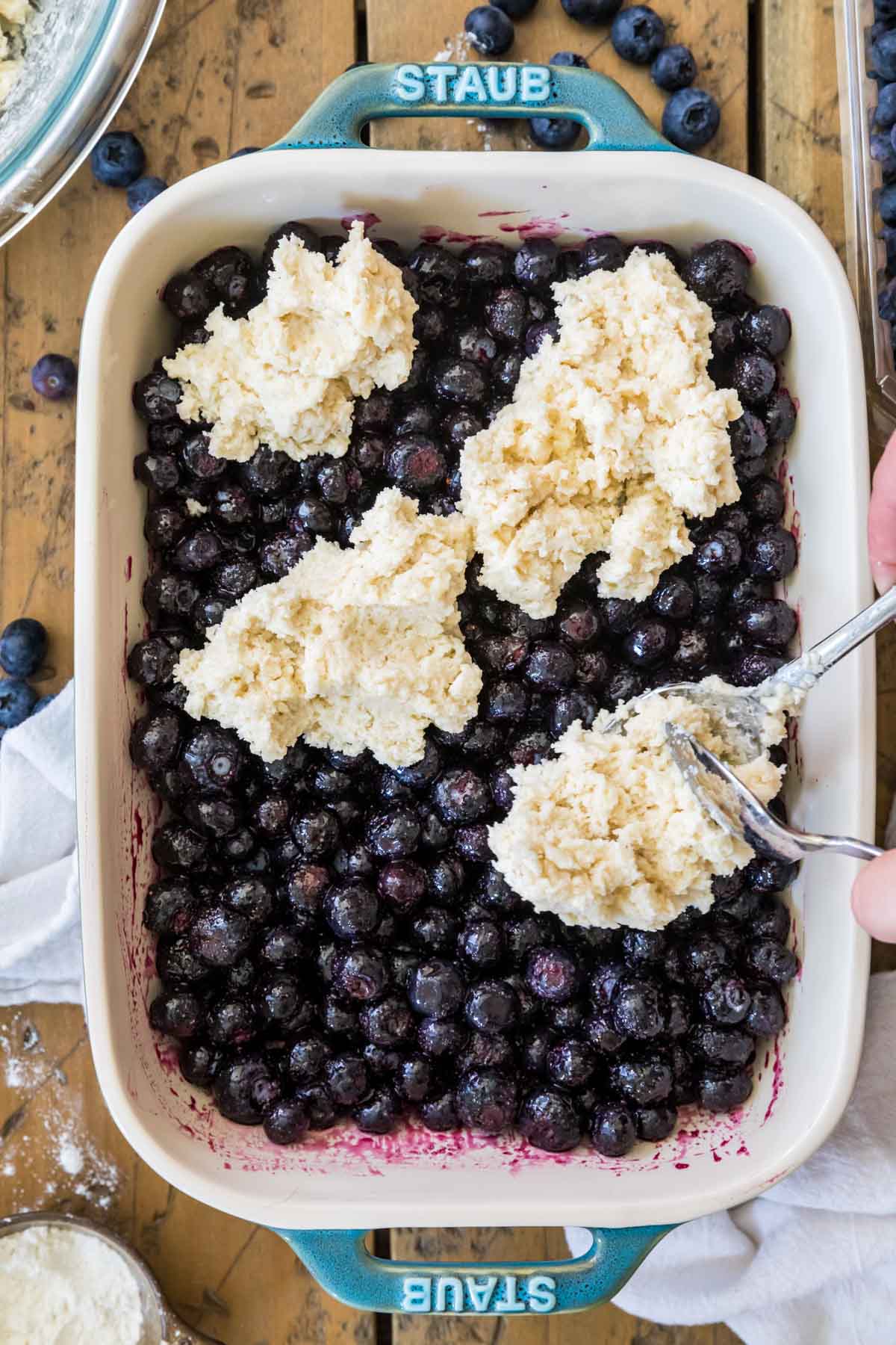 Cobbler dough being scattered over a baking dish of blueberries.