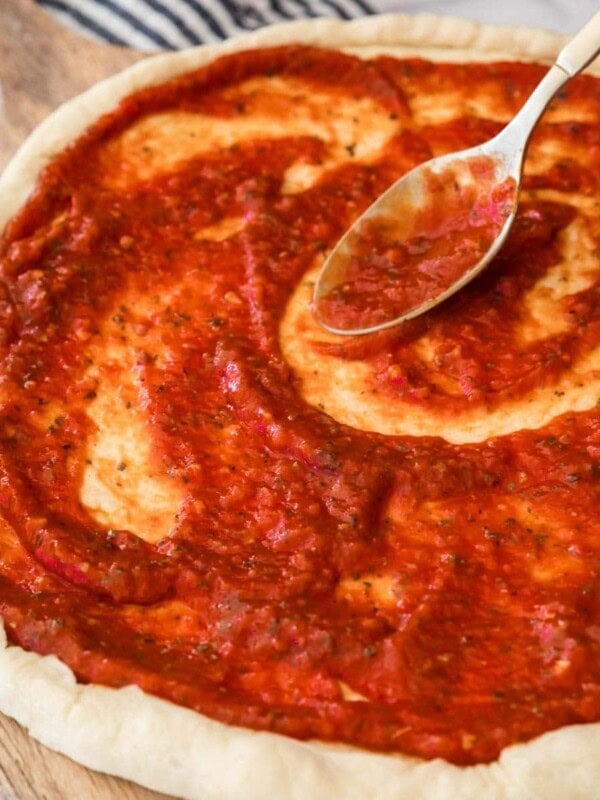 My rich red homemade pizza sauce recipe being spread onto an unbaked pizza crust.