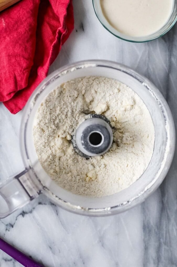Overhead view of a food processor with dry ingredients and butter pieces inside.