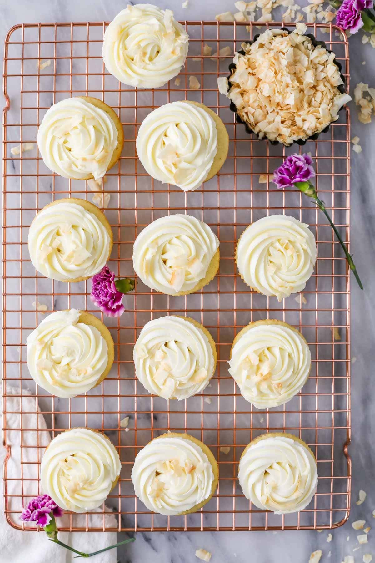 Overhead view of cupcakes topped with swirls of cream cheese frosting and toasted coconut flakes.