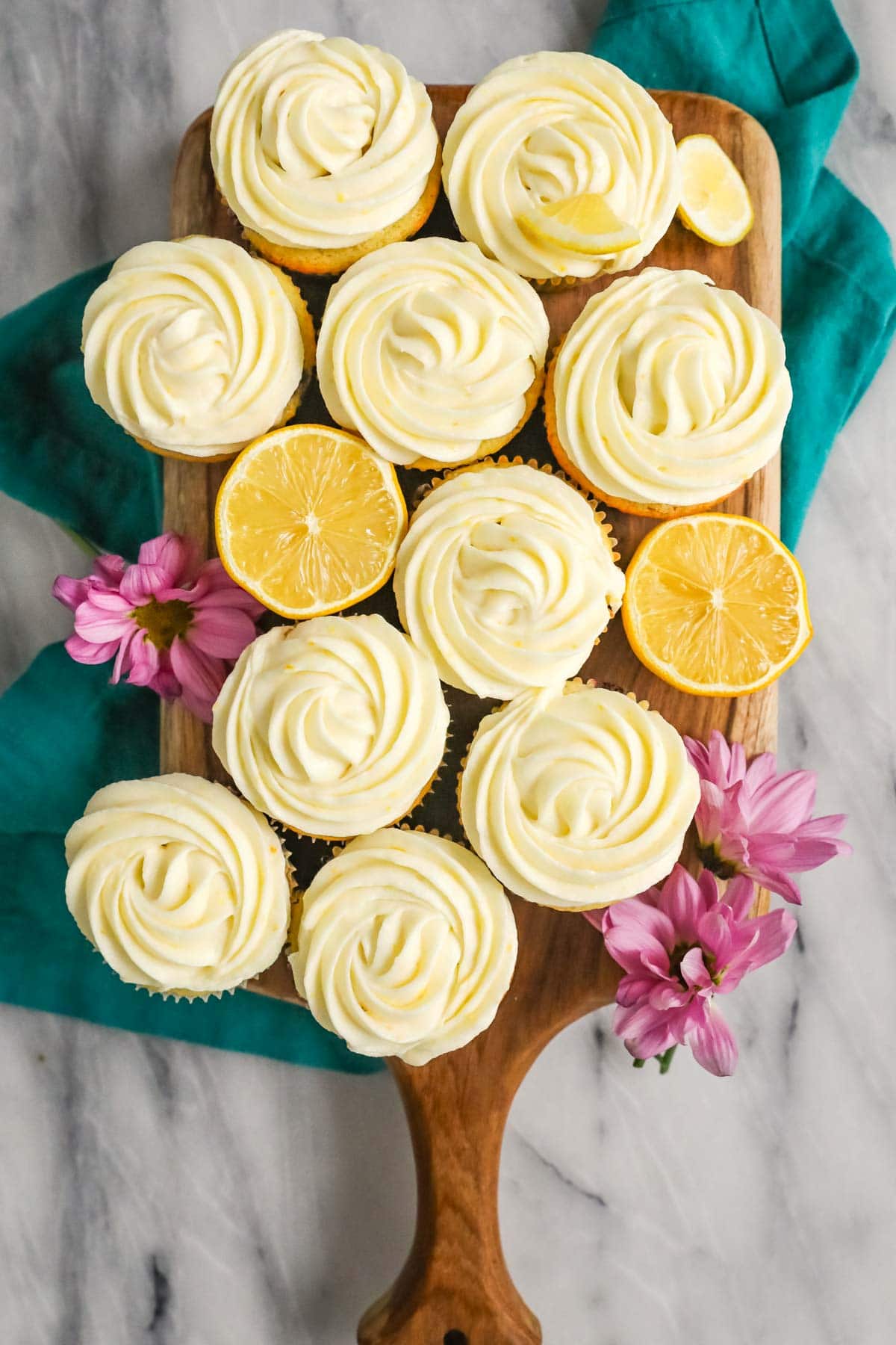 Overhead view of cupcakes frosted with lemon cream cheese frosting among lemon halves on a wood cutting board.