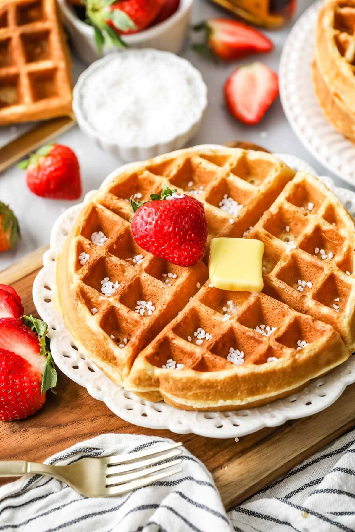 Sourdough waffle on a plate topped with a strawberry, a pat of butter, and Belgian pearl sugar.