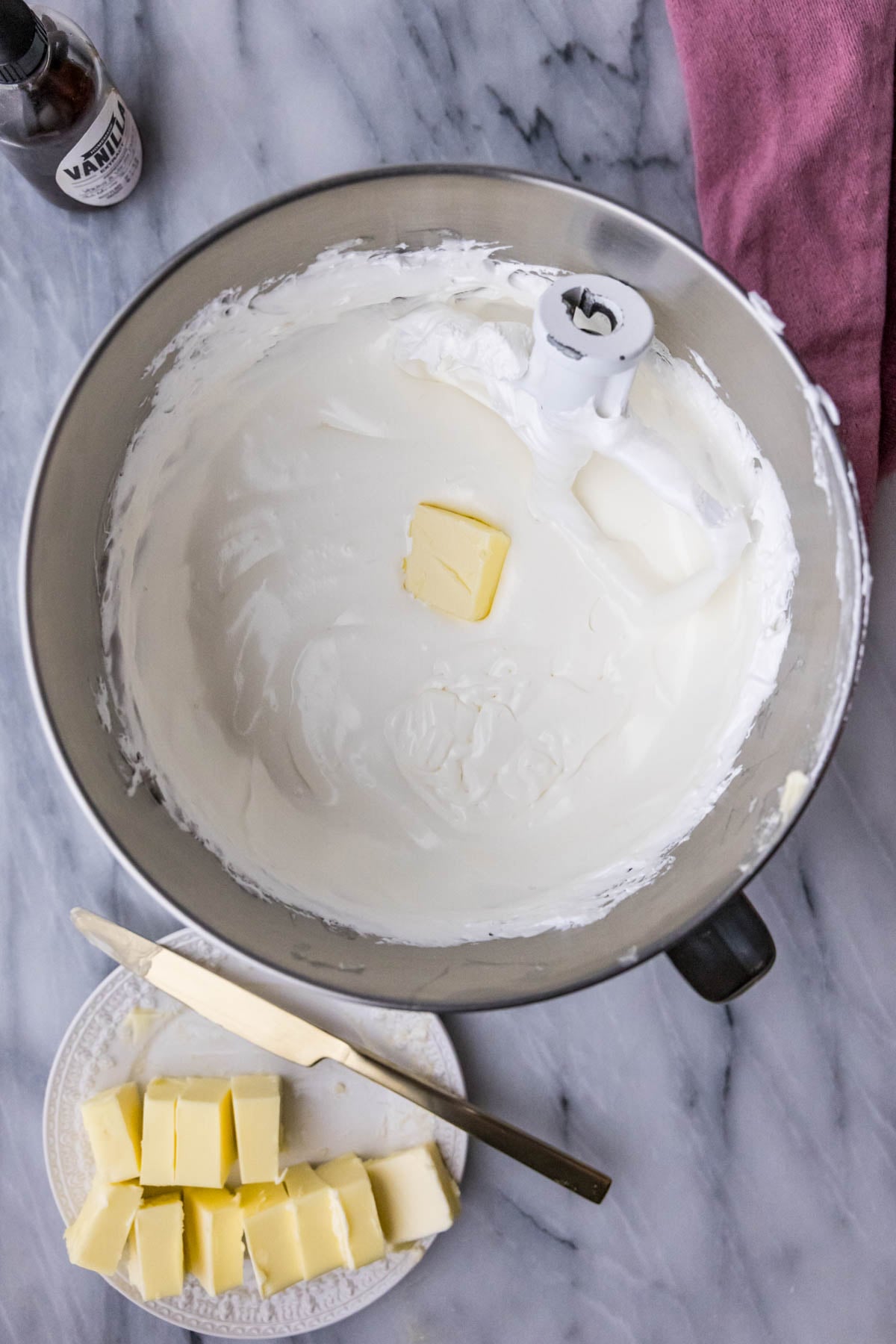 Overhead view of butter being added to meringue in a mixer bowl.